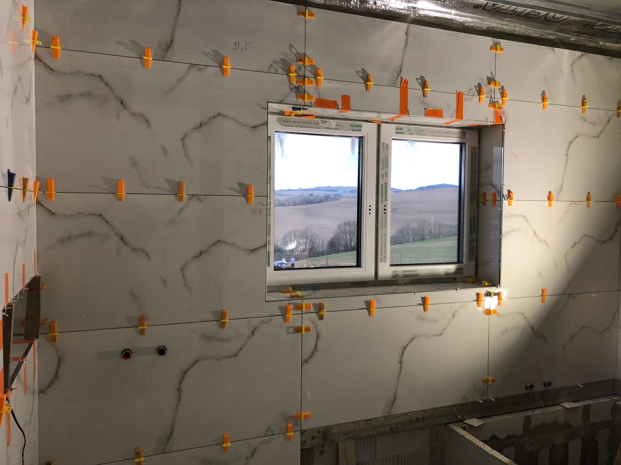 Kúpeľňa obložená veľkoformátovým mramorovým dekorom okolo okna, s nivelačnými klipmi počas kladenia